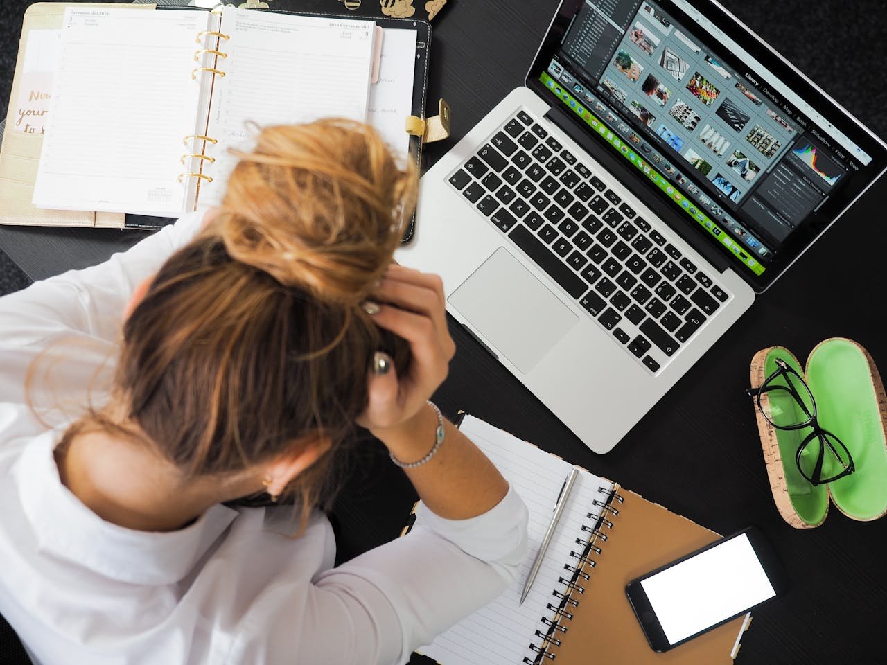 Overhead view of a stressed woman working at a desk with a laptop, phone, and notebooks.