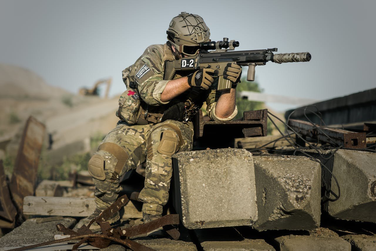 A soldier in full camouflage gear aiming a rifle while in a tactical stance outdoors.